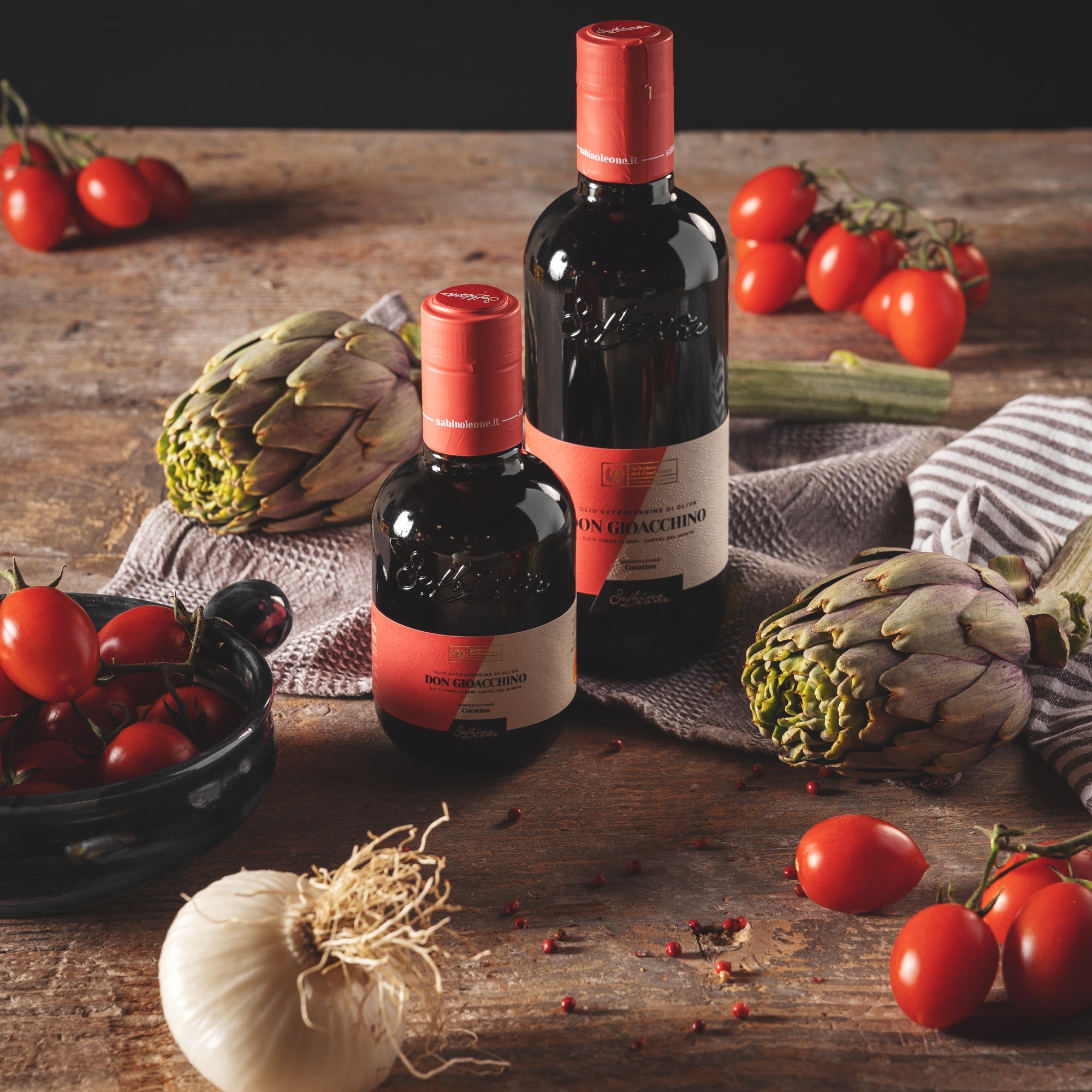 Two bottles of Sabino Leone Don Gioacchino olive oil surrounded by artichokes, tomatoes, eggplant and garlic.